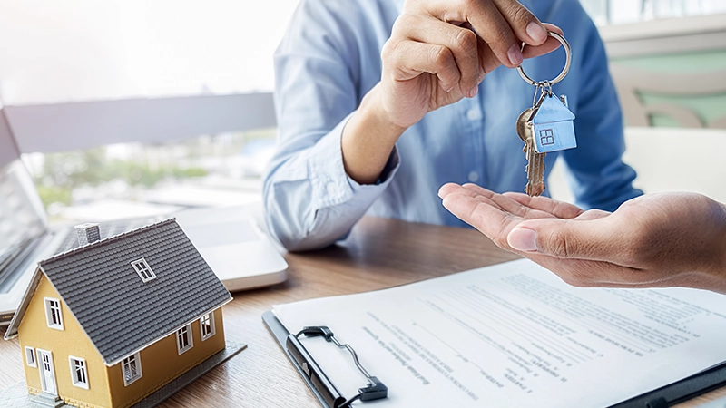 A real estate agent is handing over a set of keys, which includes a keychain shaped like a house, to a buyer.